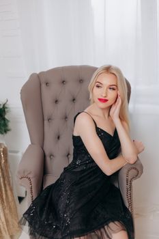 Model posing elegantly in a black dress sitting on an armchair with a white background.