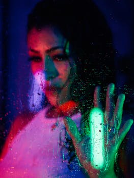Artistic portrait of a woman behind wet glass with dramatic green and pink lighting in a moody setting.