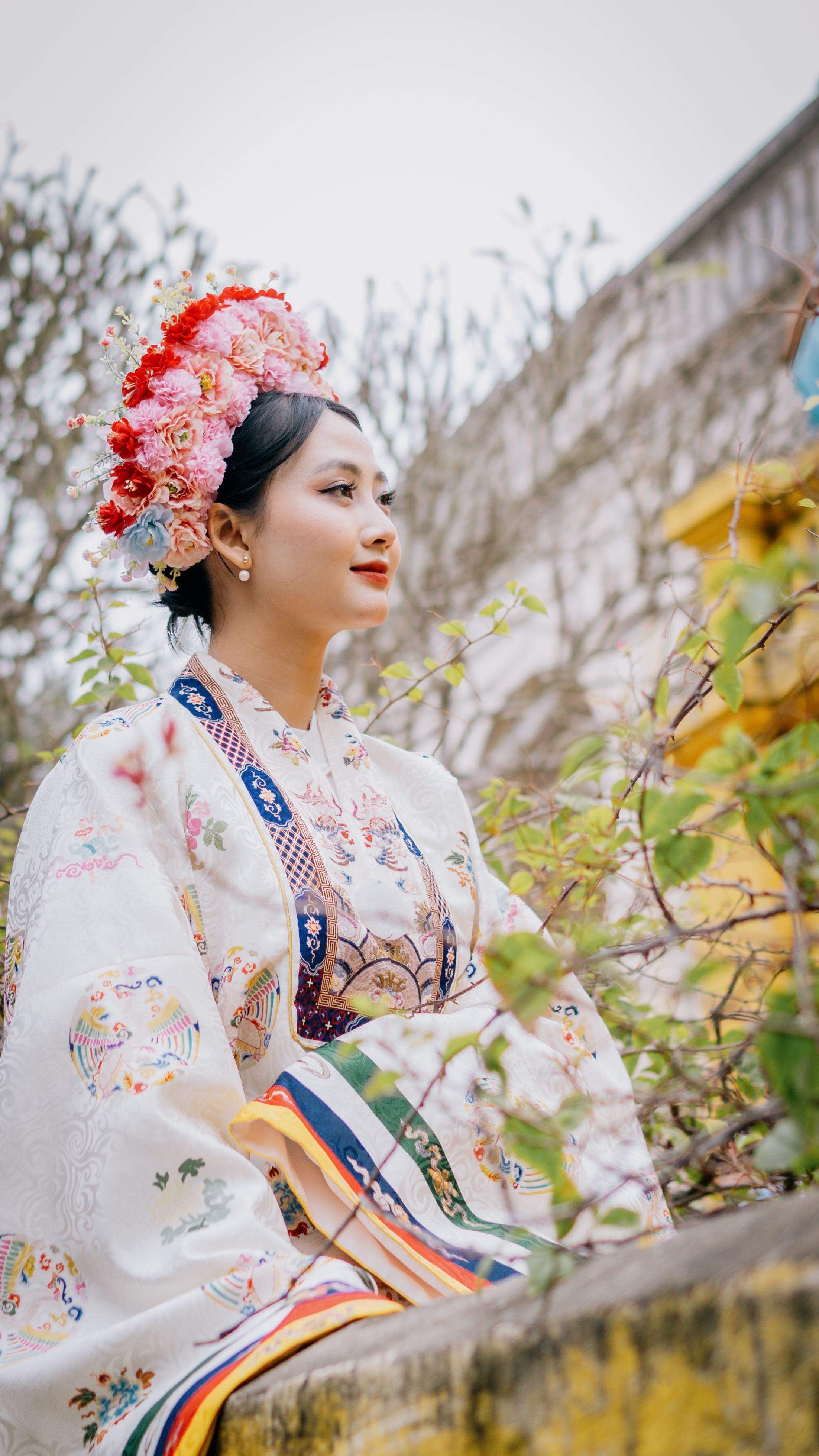 Asian bride in a traditional gown and floral headpiece posed outdoors in a serene setting.