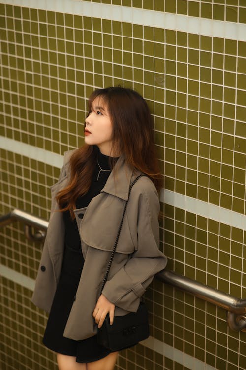 A woman in a black dress and trench coat leaning against a wall