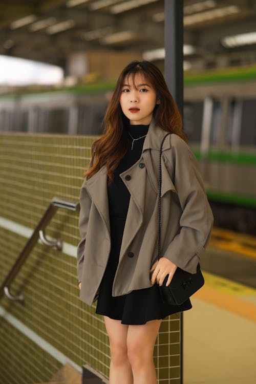 A woman in a black dress and a gray coat standing at a train station
