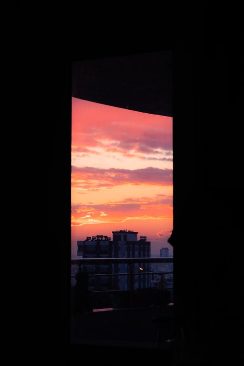 Free Silhouette of city buildings against a vibrant sunset viewed through a window. Stock Photo