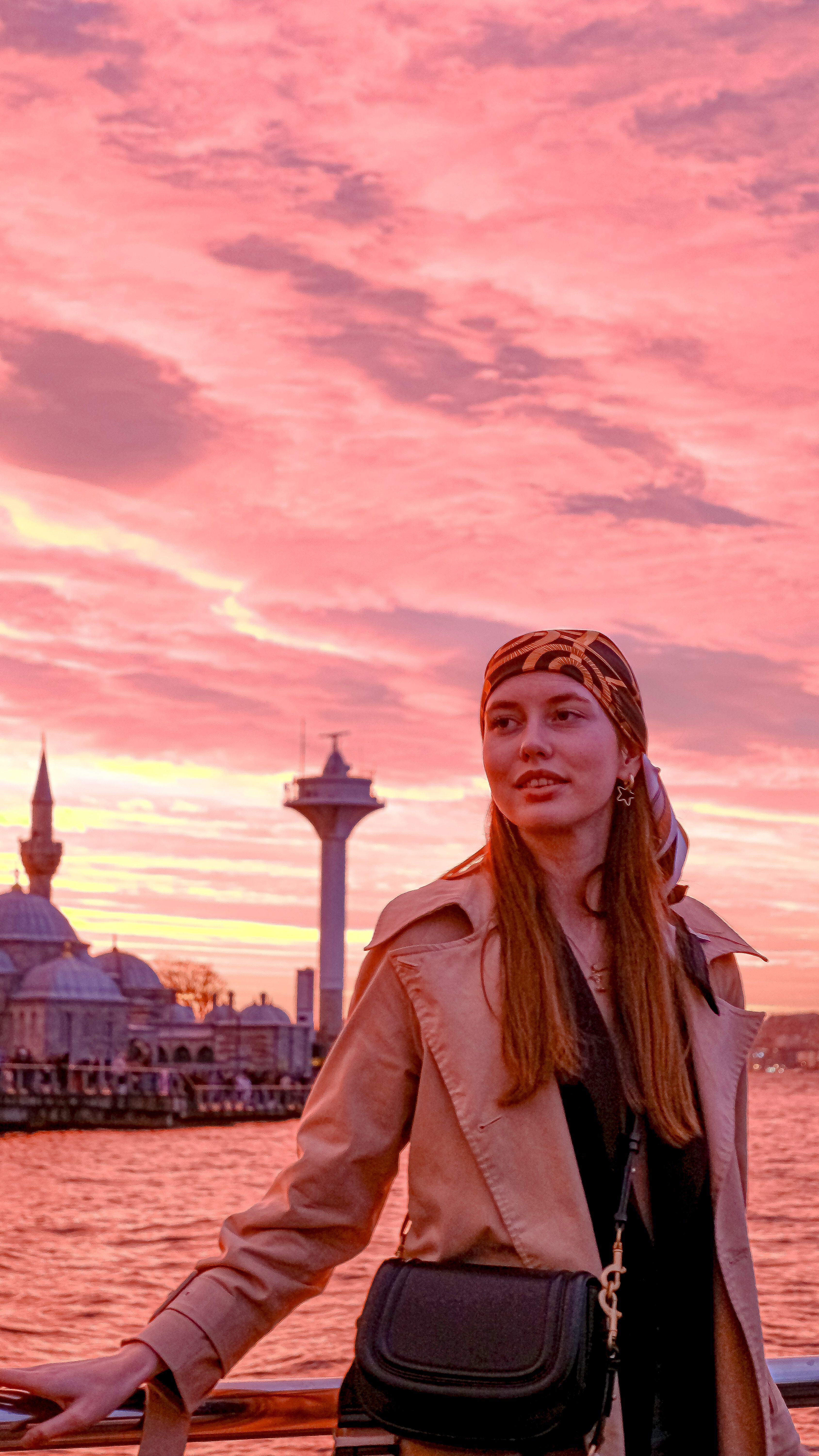 A woman poses by the Istanbul waterfront at sunset, with iconic landmarks in the background.