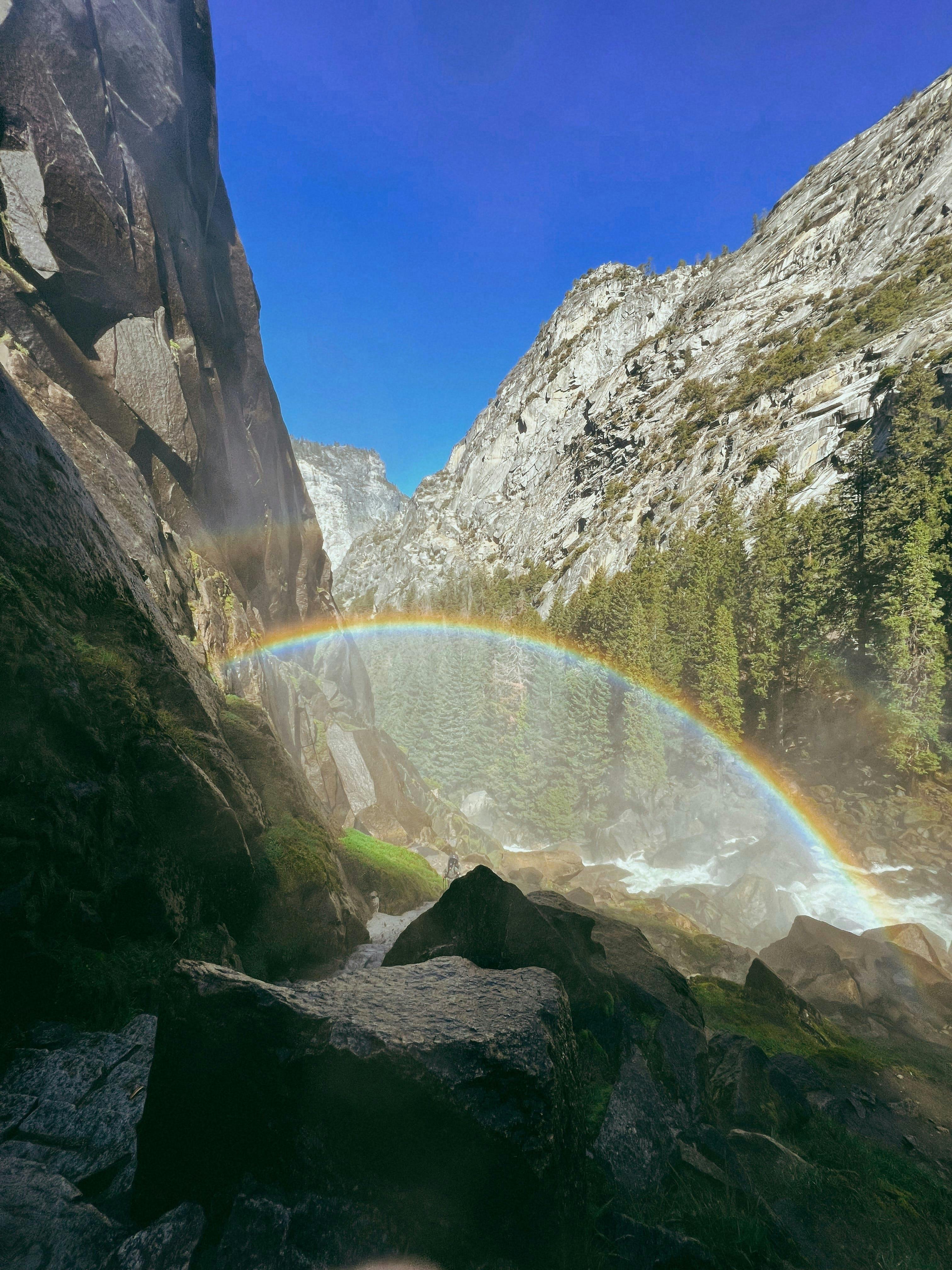 Vibrant rainbow arching over a rocky mountain valley and lush forest in California.