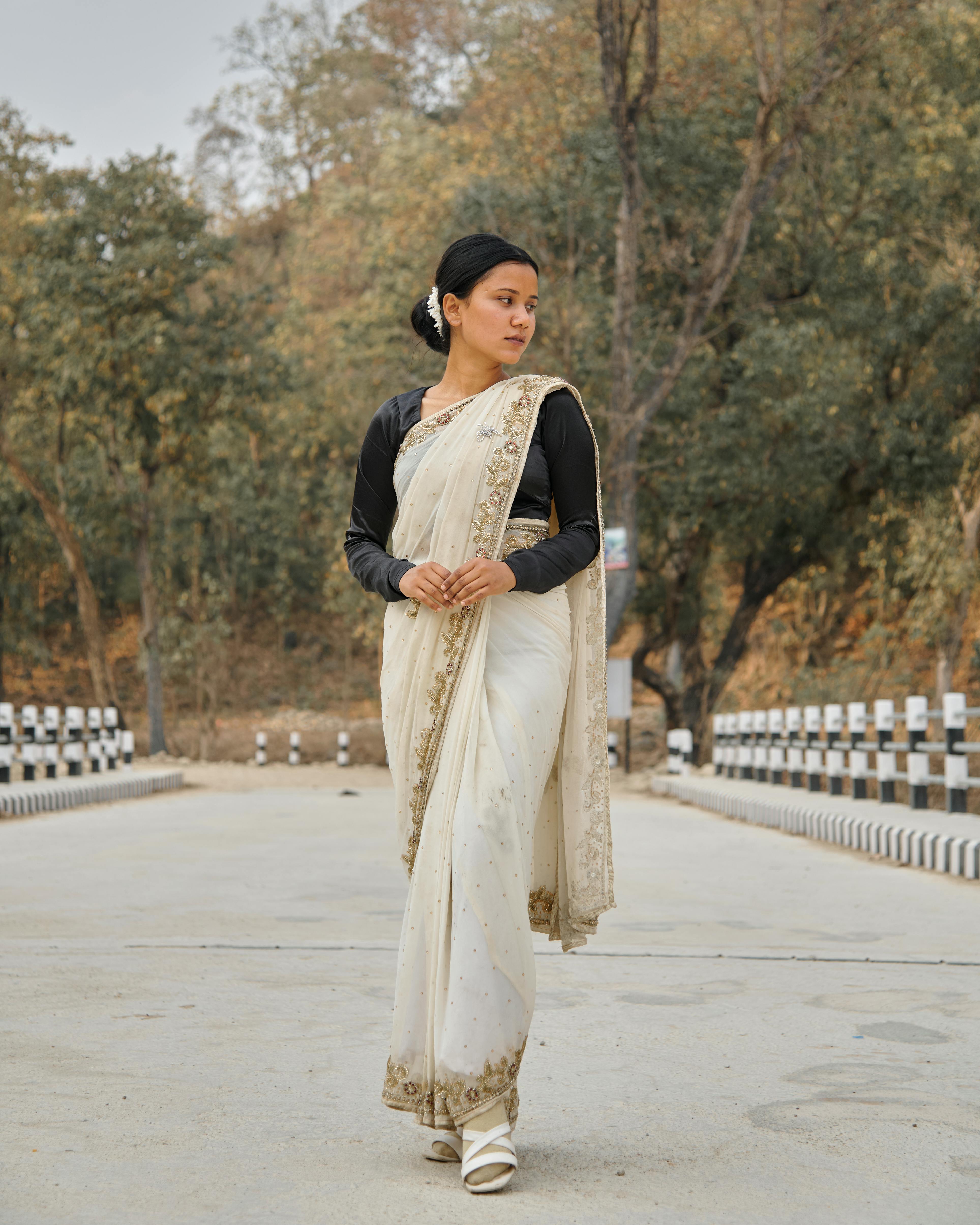 A woman in a saree gracefully walks through a serene park setting, wearing heels and expressing elegance.