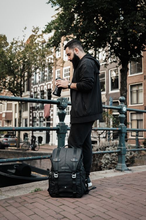 Free Bearded man with backpack and camera standing on a canal bridge, urban setting. Stock Photo