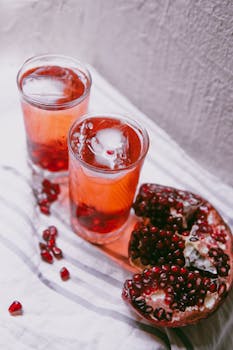 Two glasses of pomegranate juice with ice cubes and fresh fruit on a table.