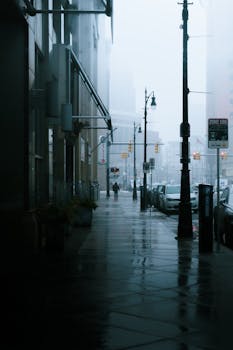 A foggy street scene in Detroit featuring wet sidewalks and streetlights reflecting on a rainy day.