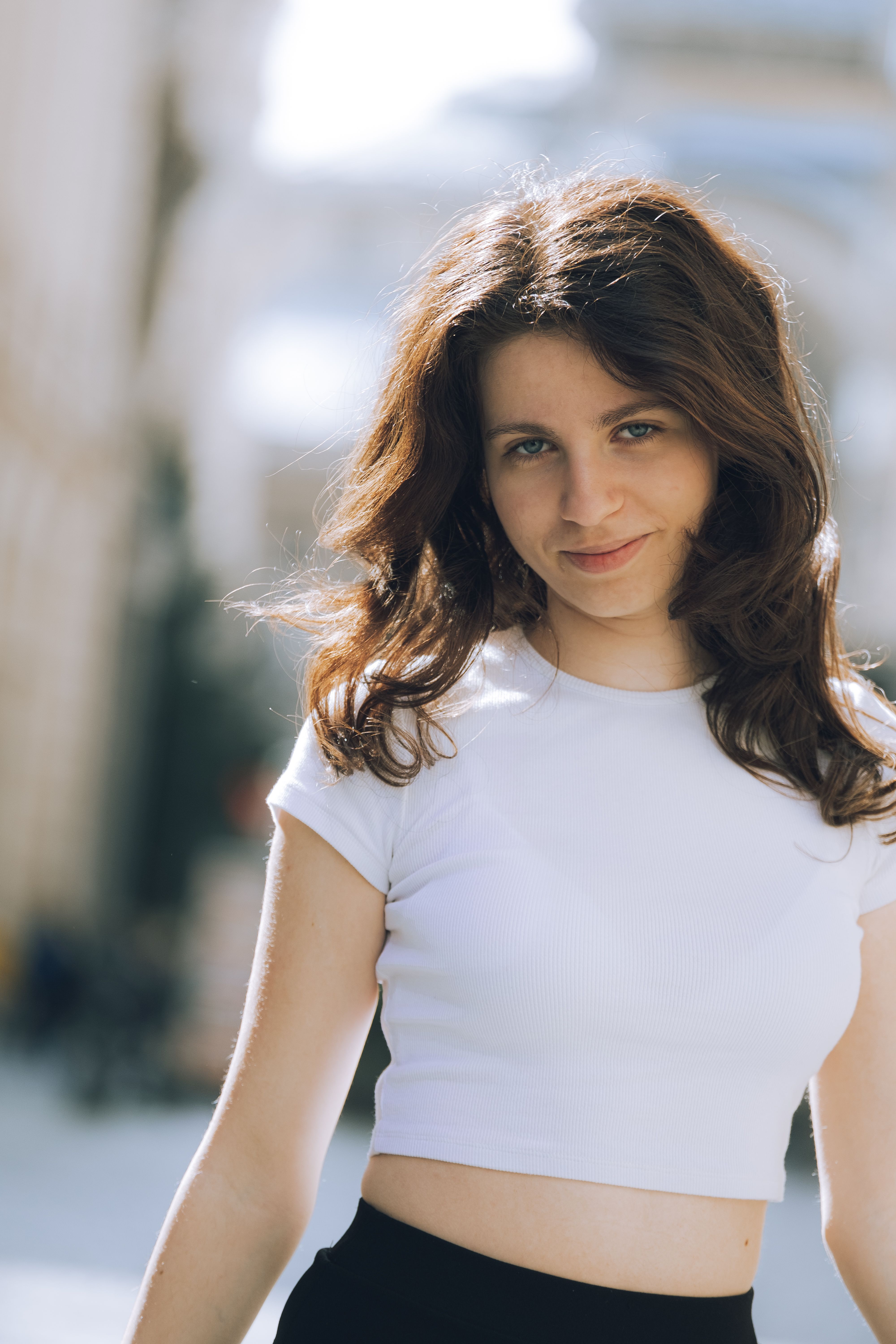 Confident young woman with brunette hair smiling in casual white crop top, standing outdoors.