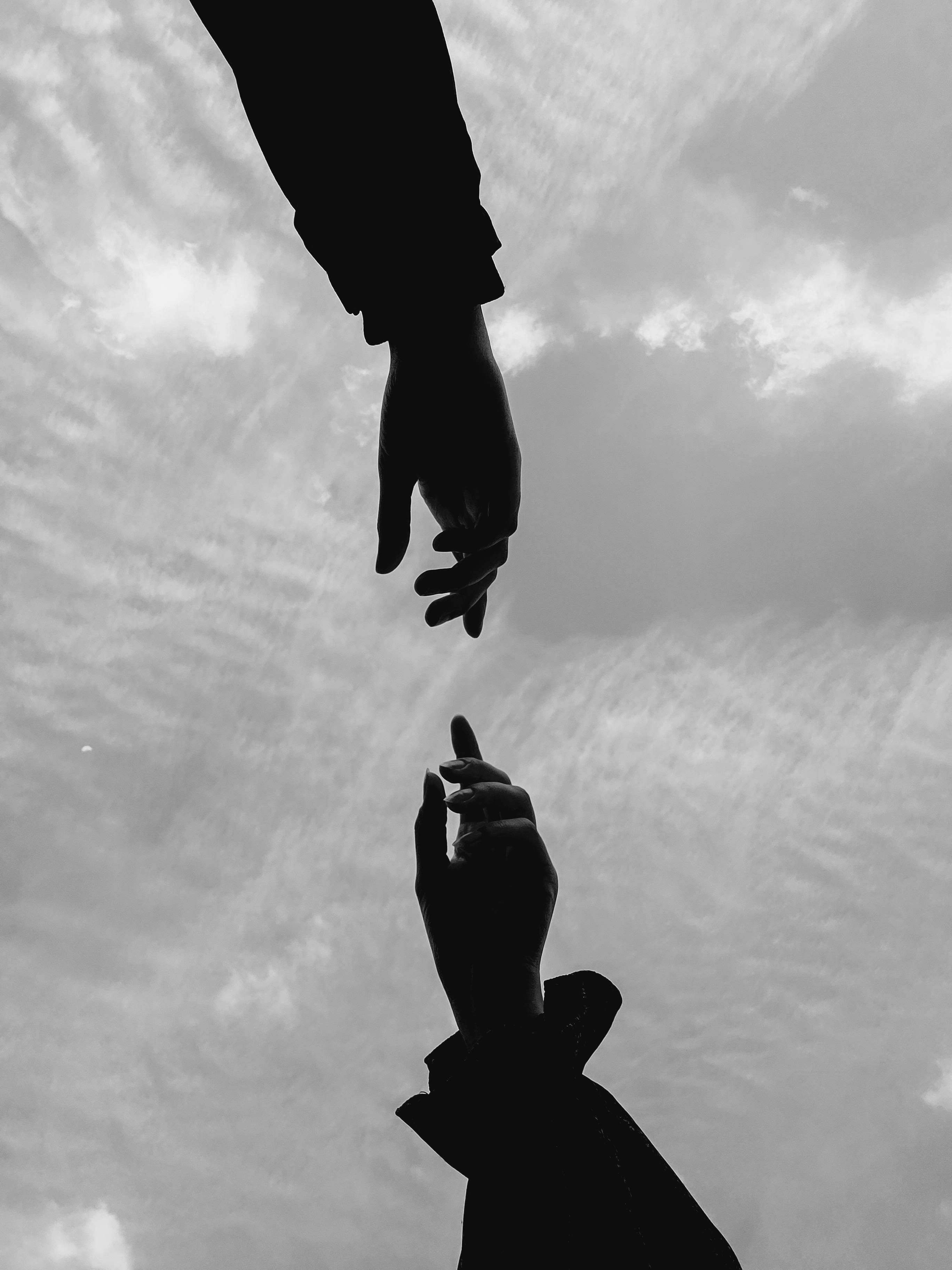 A dramatic black and white photo capturing two hands reaching towards each other against a clouded sky.