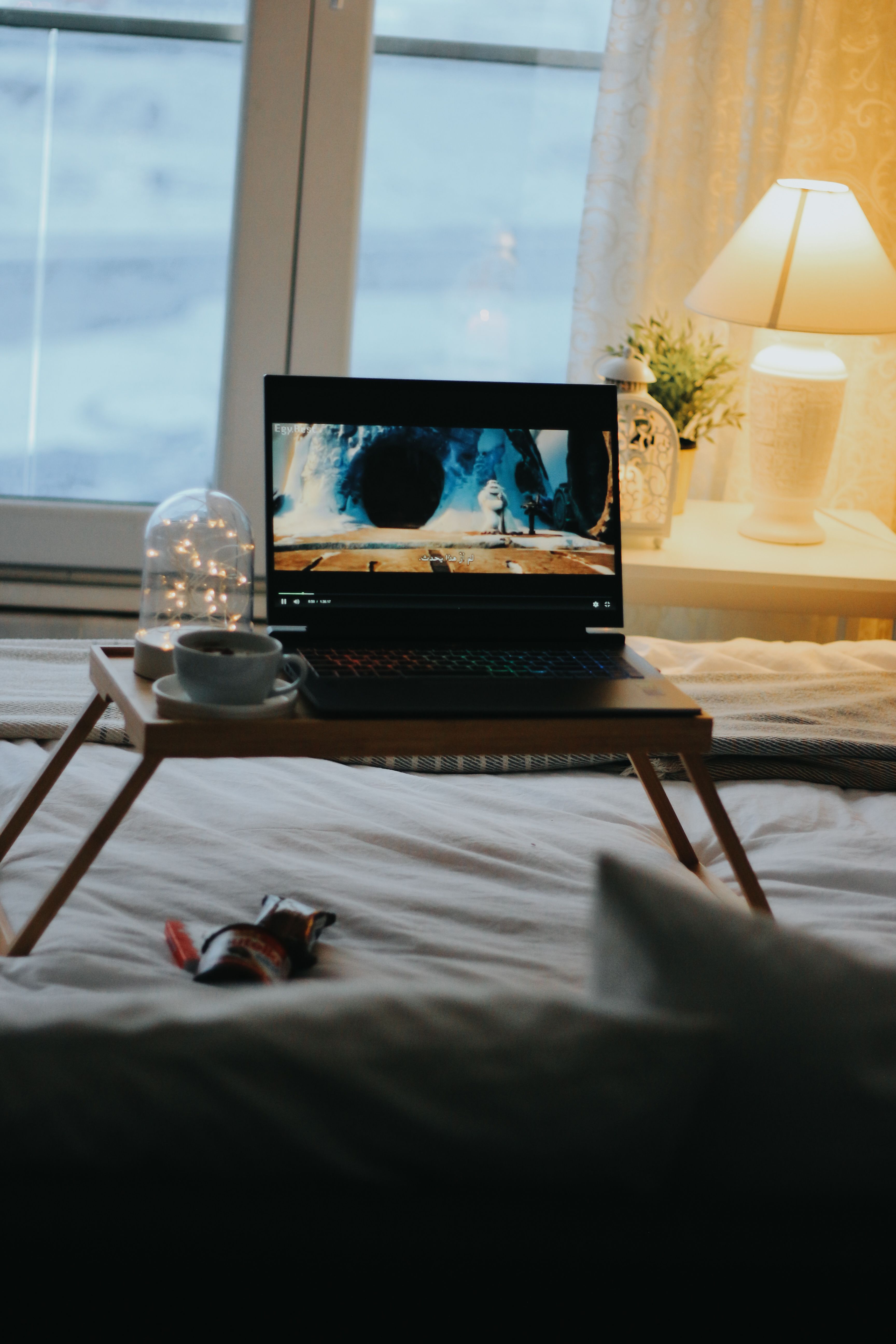 A cozy bedroom scene featuring a laptop on a bed tray near a warm lamp, ideal for relaxation.