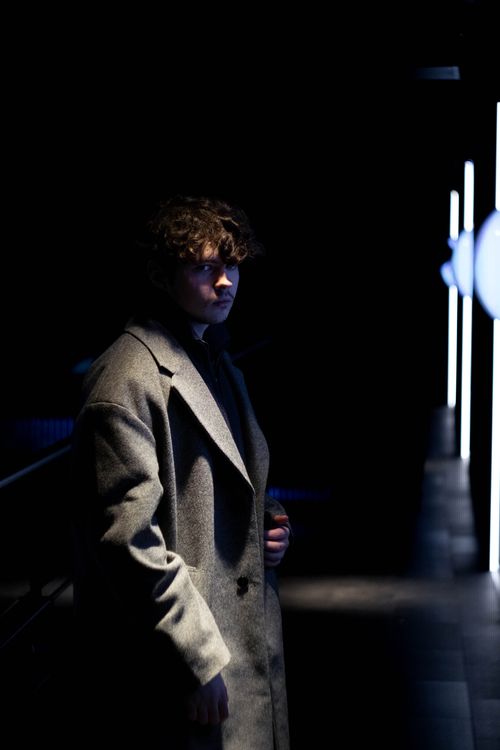 Free A young man with curly hair stands in a dark, illuminated urban area at night wearing a gray coat. Stock Photo