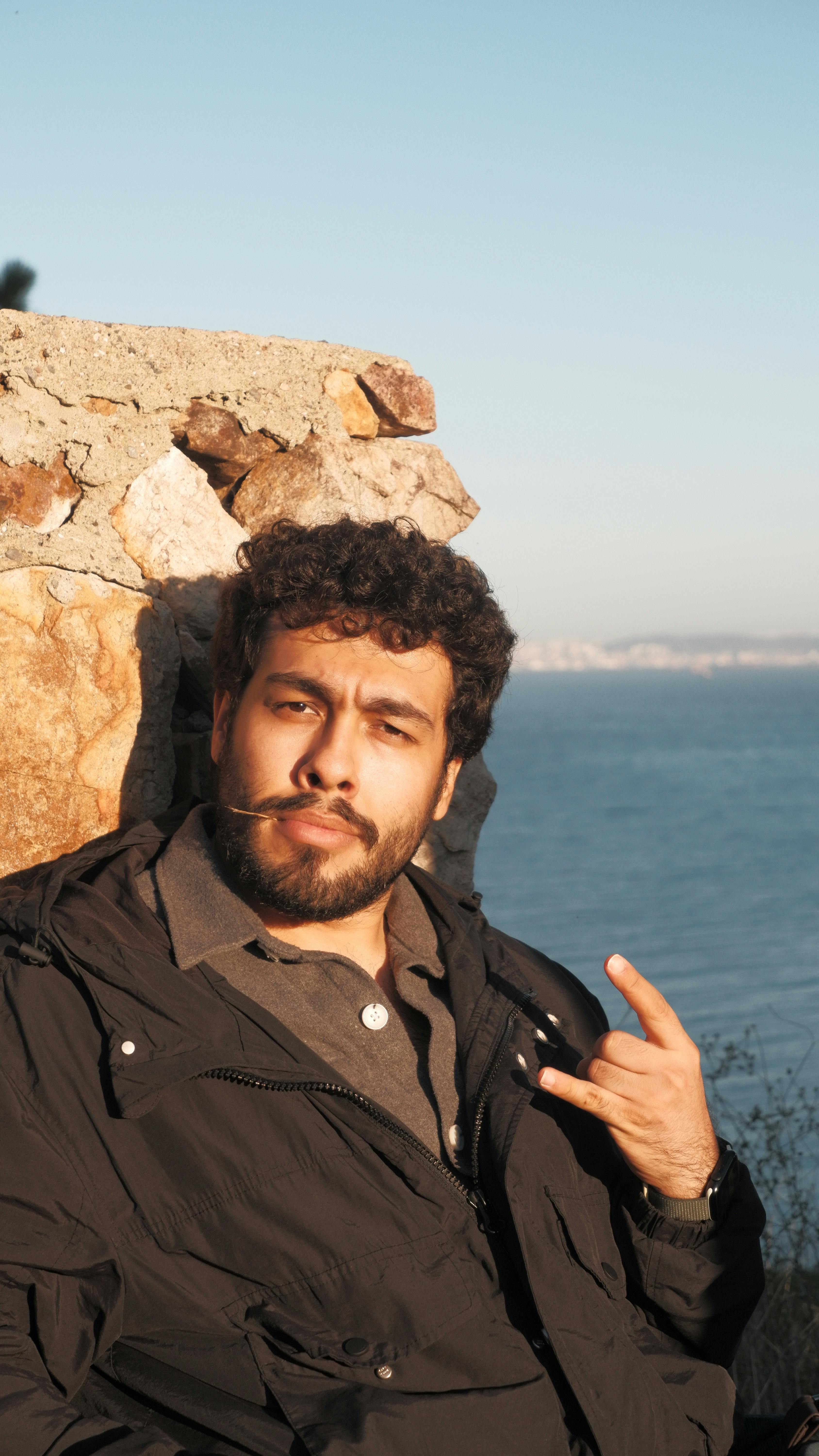 A man with a beard and mustache sits casually by the seaside in bright sunlight, exuding relaxation.