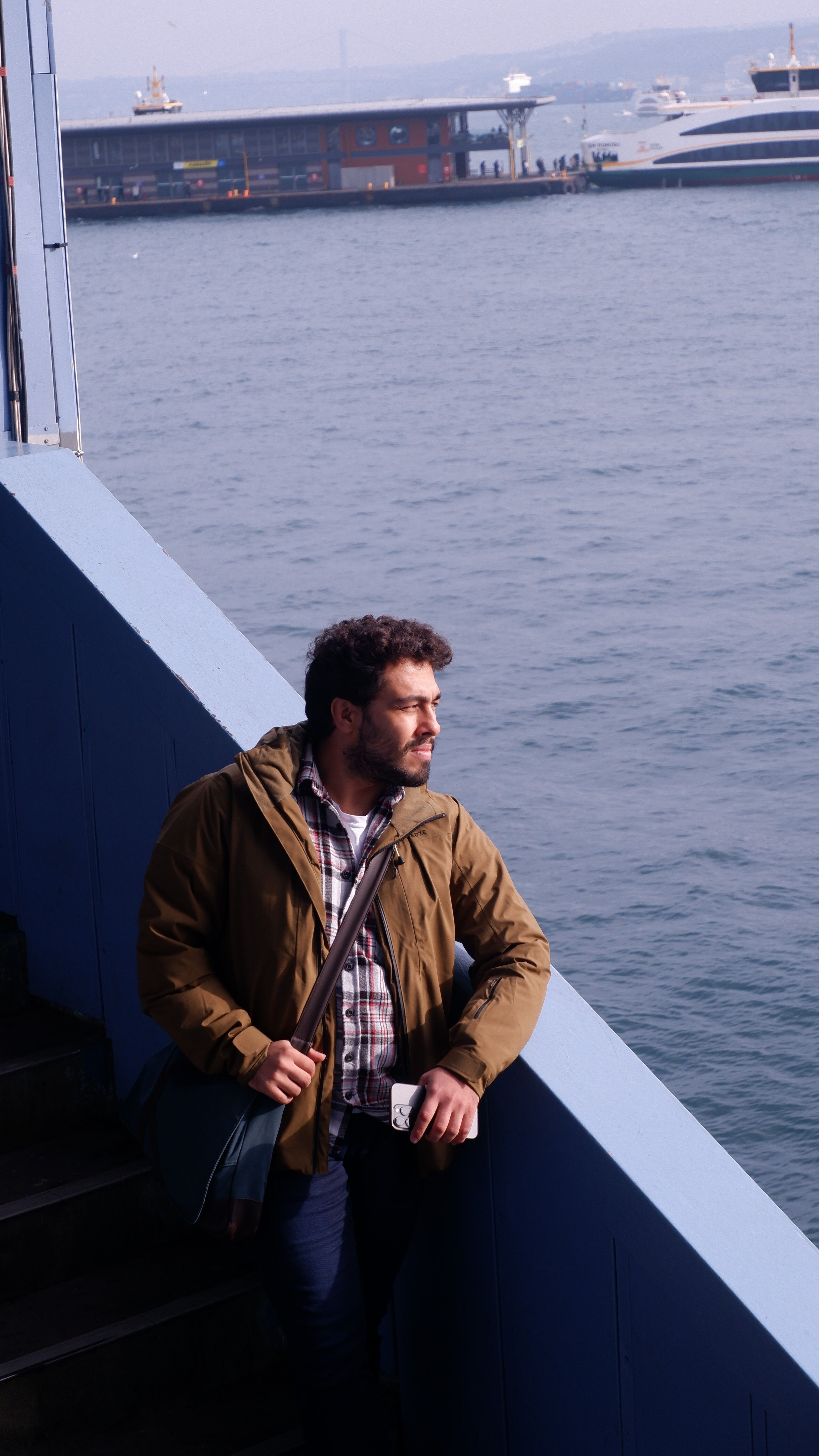 Man in jacket gazing at ocean from waterfront in urban setting, capturing a serene moment of reflection.