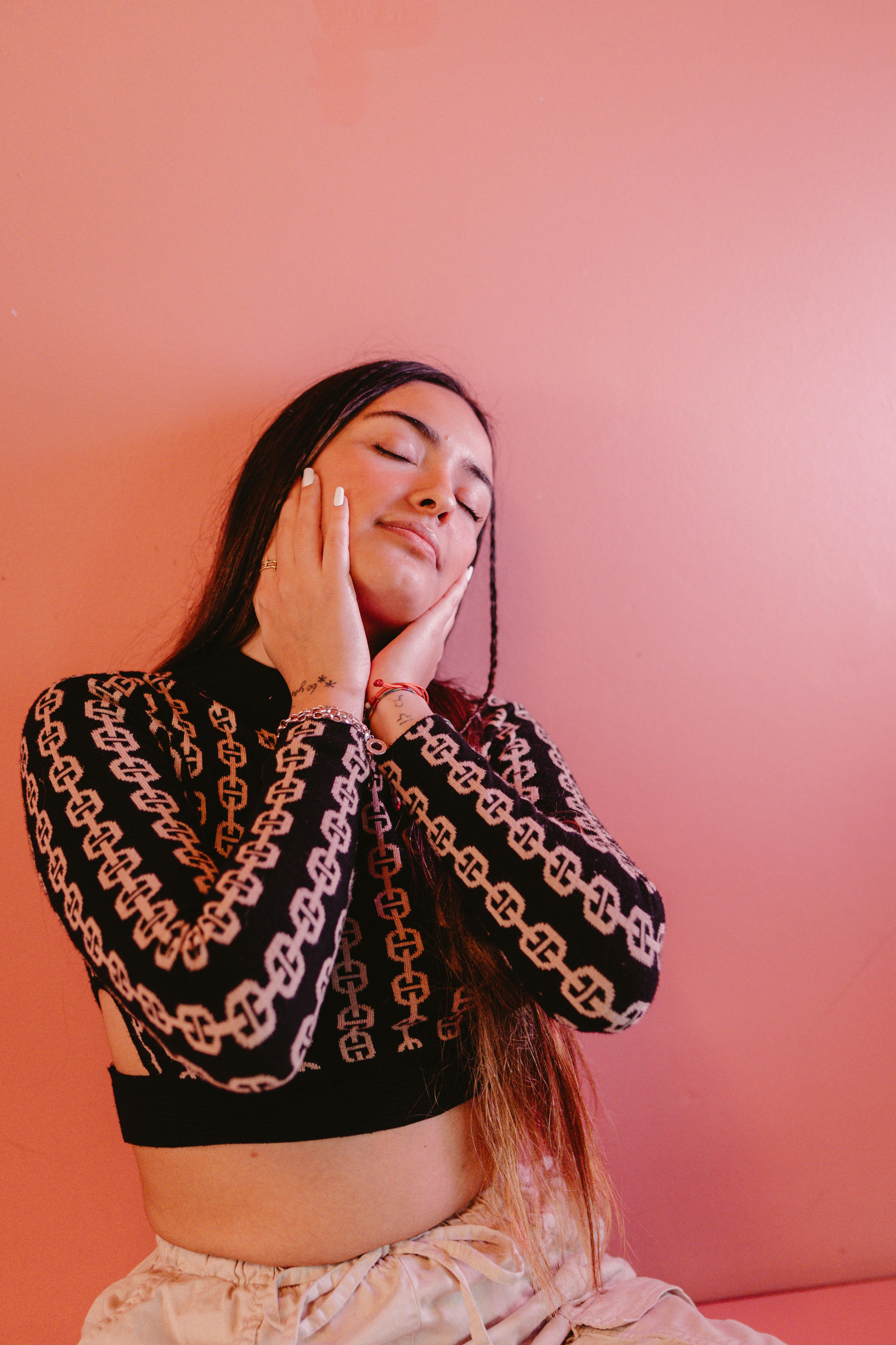 Portrait of a calm woman with long hair, eyes closed against a pink wall, embodying tranquility and style.