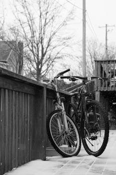 A black and white winter scene featuring parked bicycles against a railing in an urban setting.