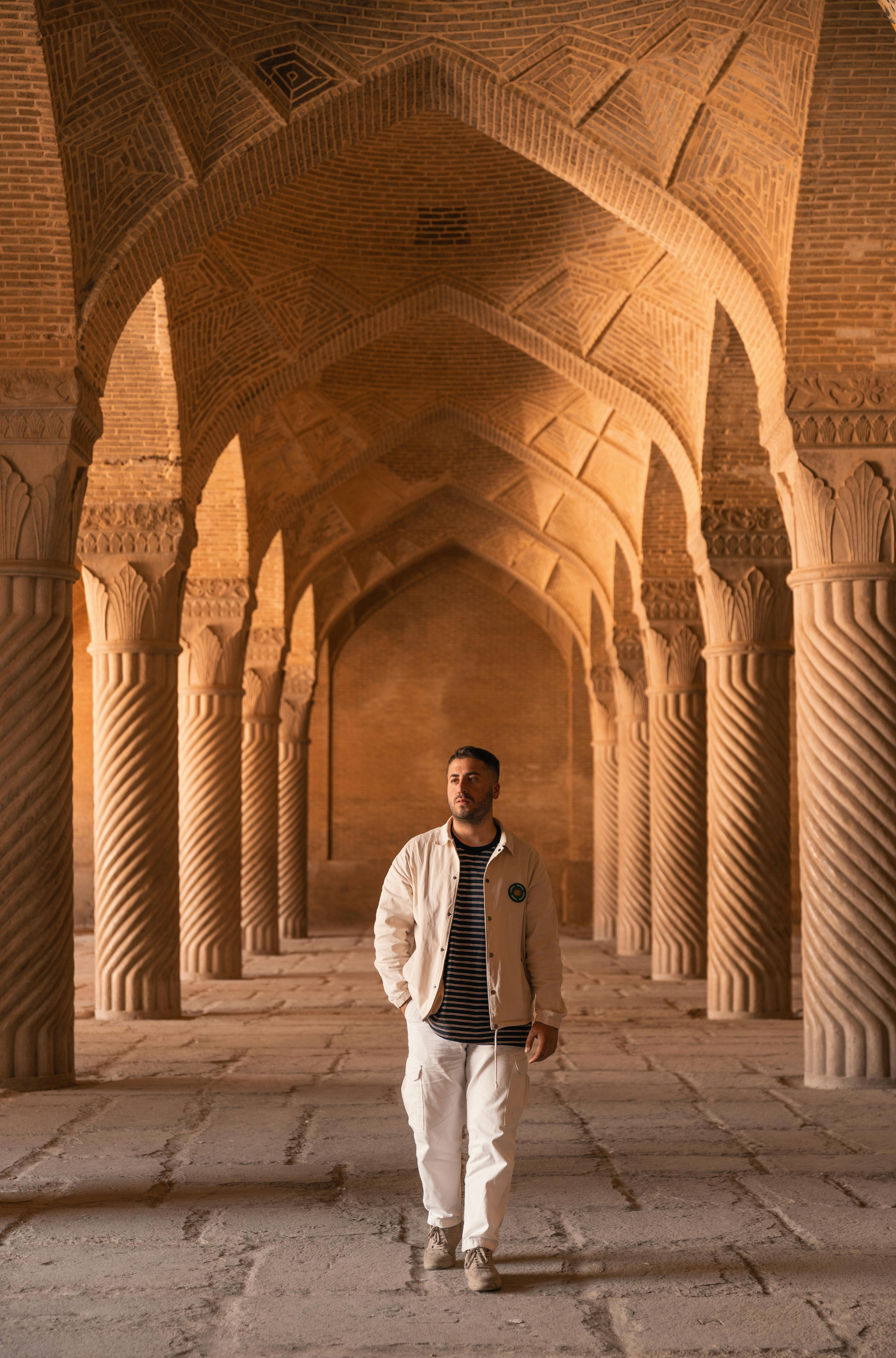 Young man in white jacket walking through a historical colonnade, showcasing architectural beauty.