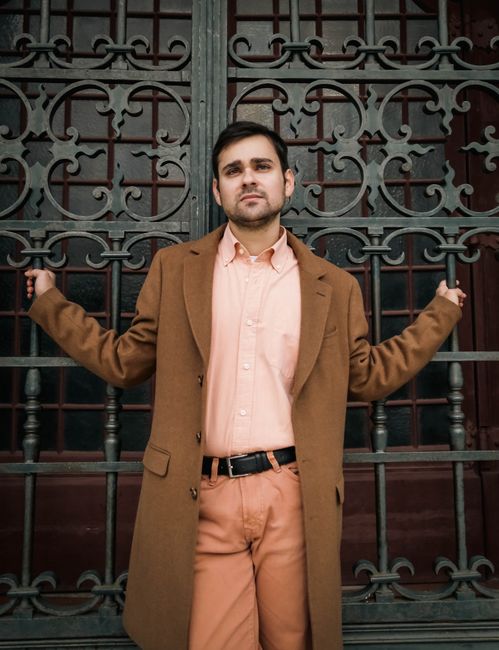 Stylish urban portrait of a man standing in front of an ornate wrought iron gate wearing a brown coat.