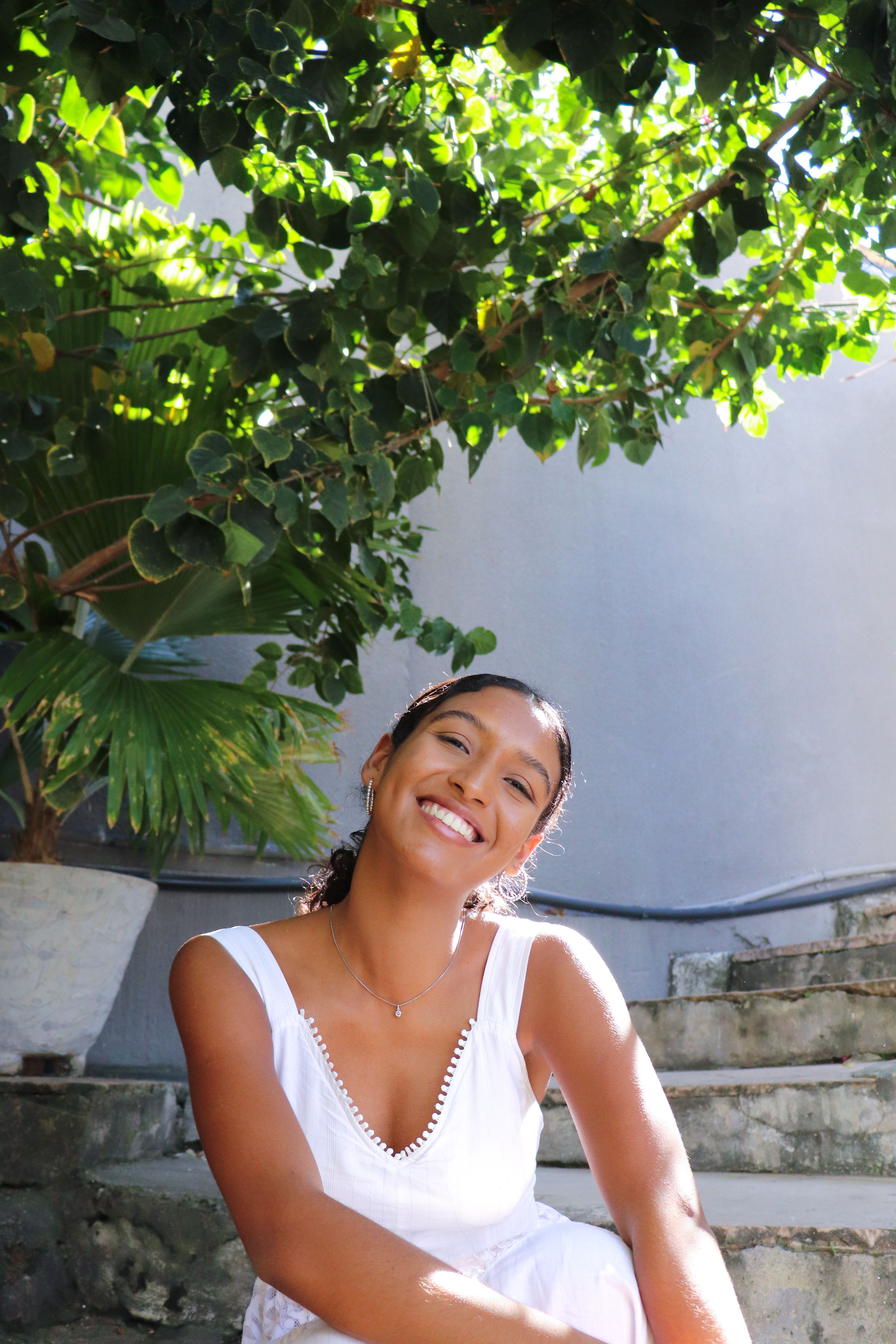 Happy woman in white dress sits outdoors under green foliage, smiling brightly.