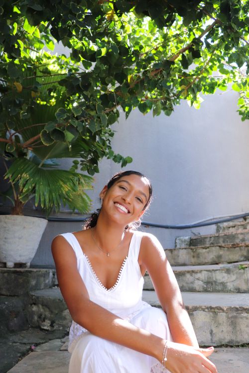 Free A young woman joyfully sitting on stairs under lush greenery, wearing a white dress on a sunny day. Stock Photo