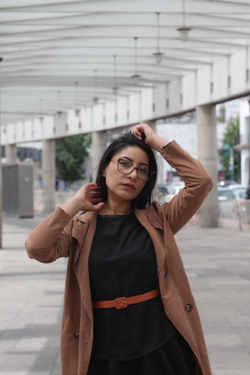 Free Confident woman with glasses posing outdoors in a fashionable brown coat in a modern urban environment. Stock Photo