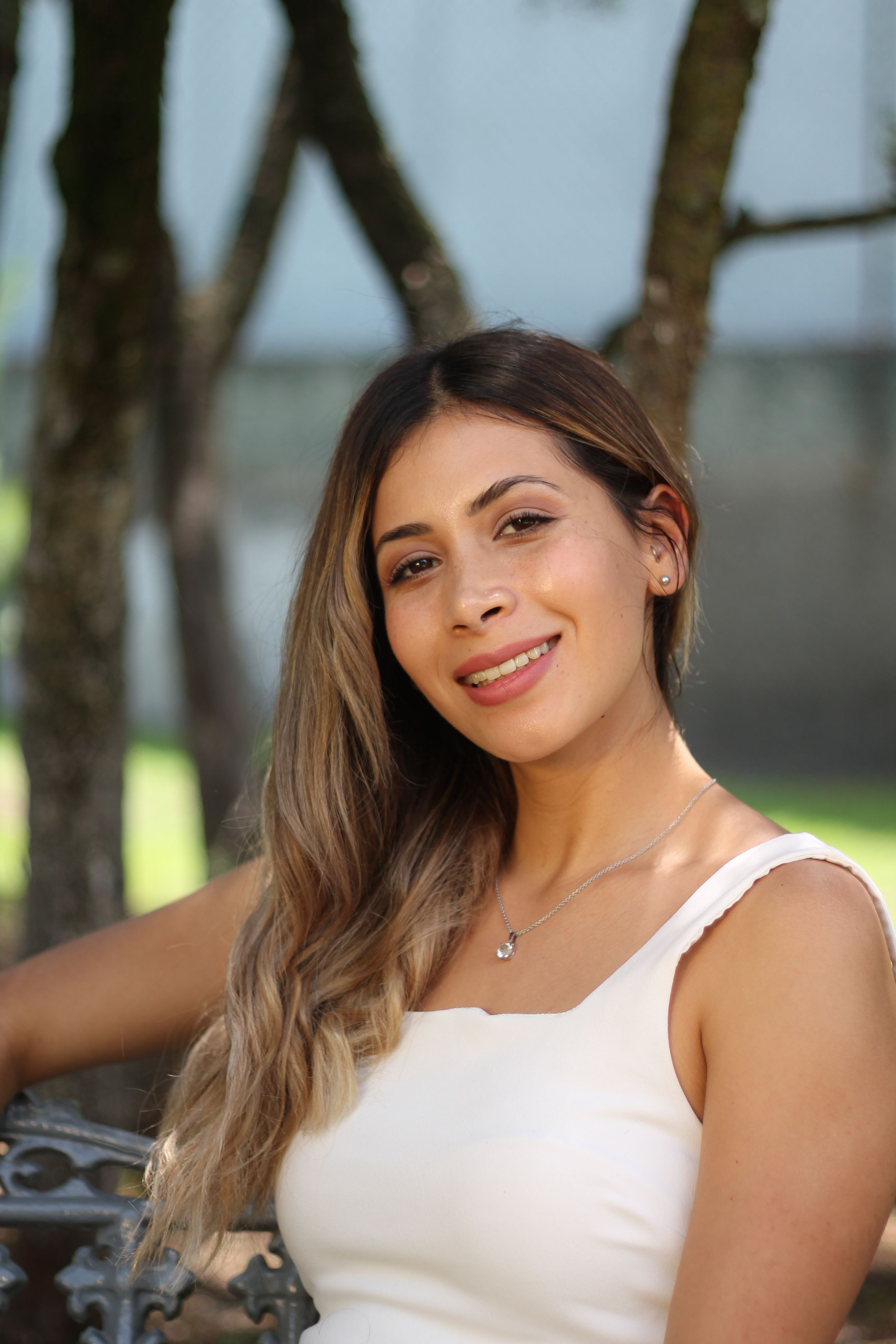 Portrait of a smiling woman sitting on a park bench with trees in the background.