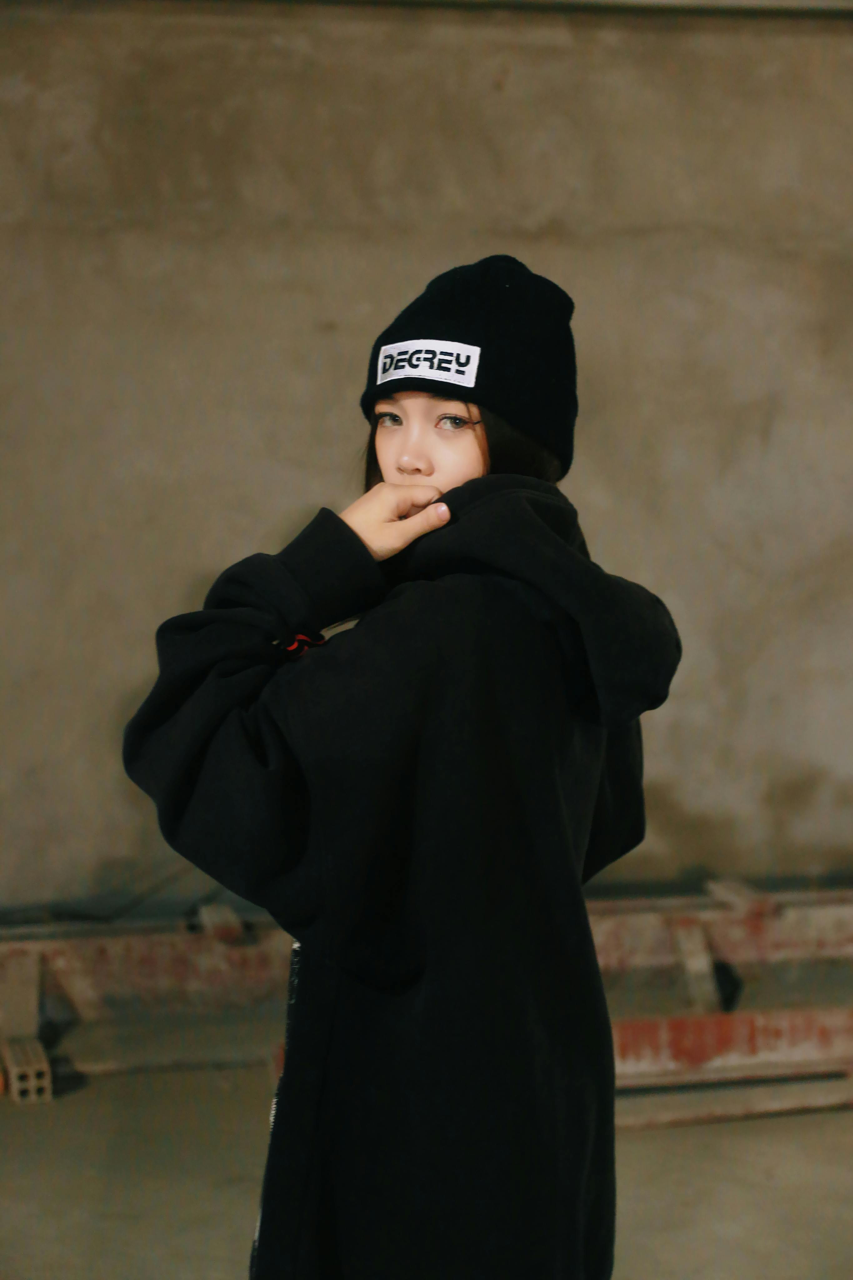 Young woman in a black hoodie and beanie, posing indoors with a moody expression.