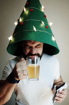 Man enjoying beer with a quirky Christmas tree hat indoors.