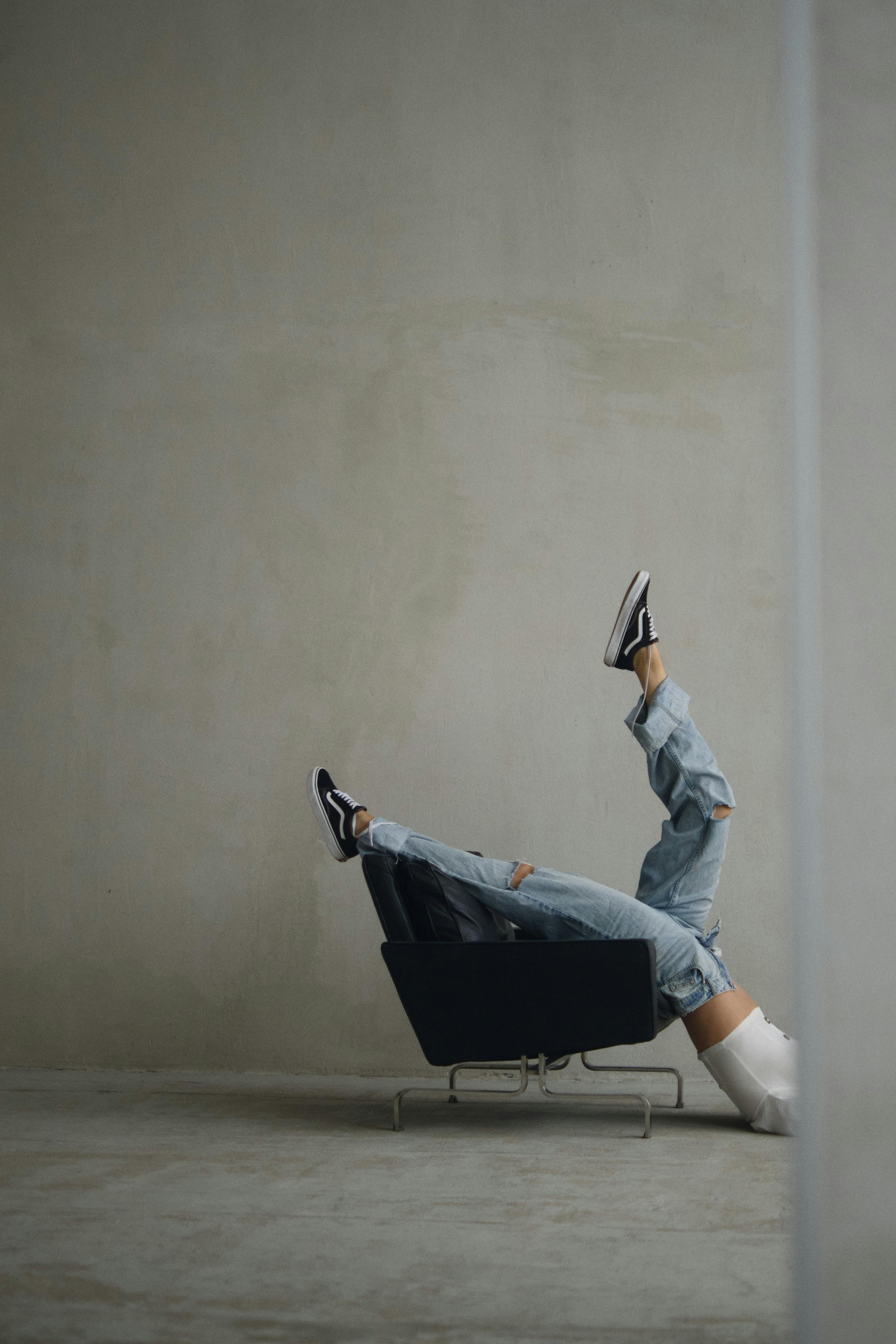 Artistic pose of woman in jeans and sneakers with legs raised on modern chair against minimalistic wall.