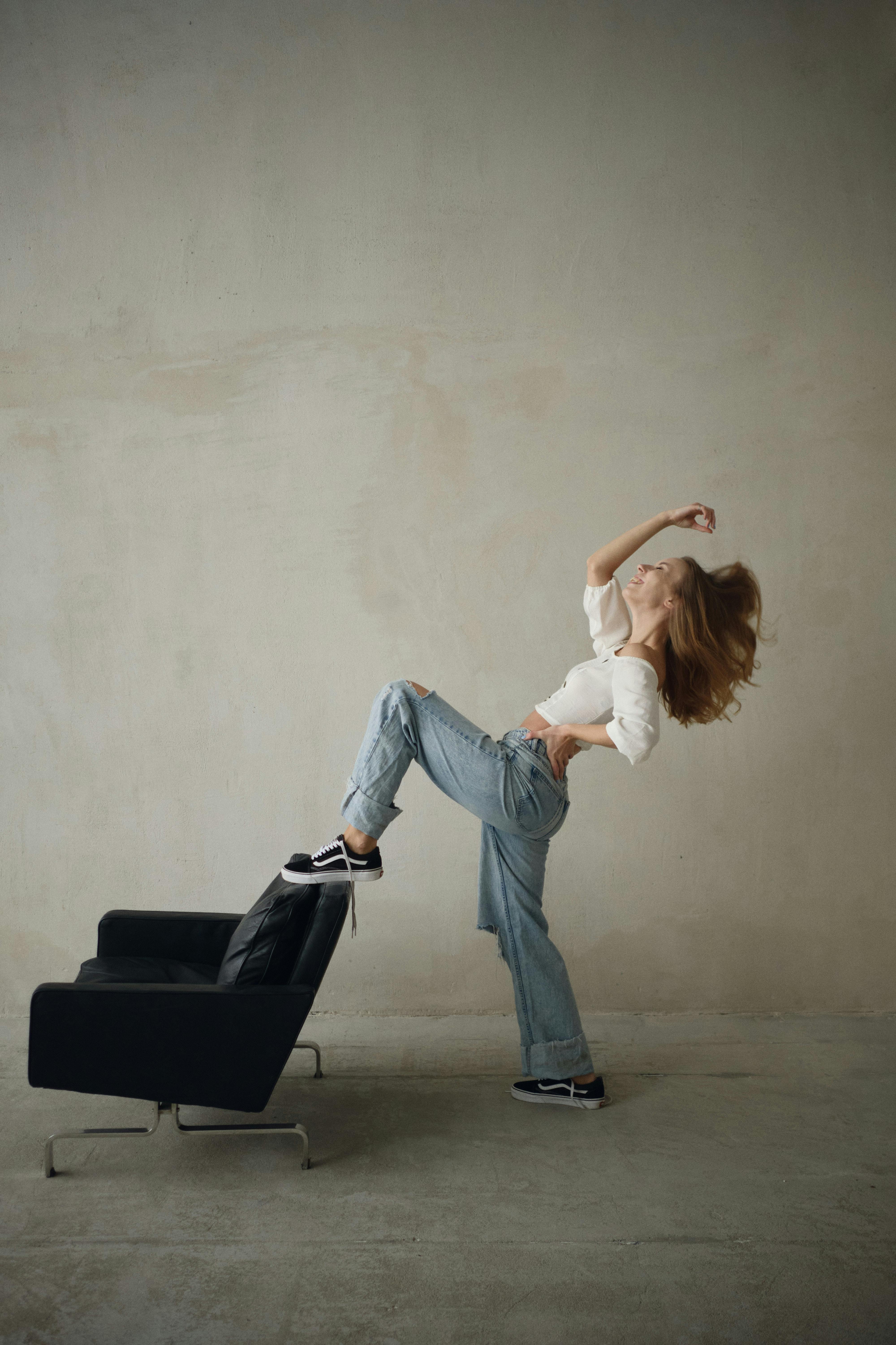 Young woman in casual attire posing dynamically on an armchair in a studio setting.