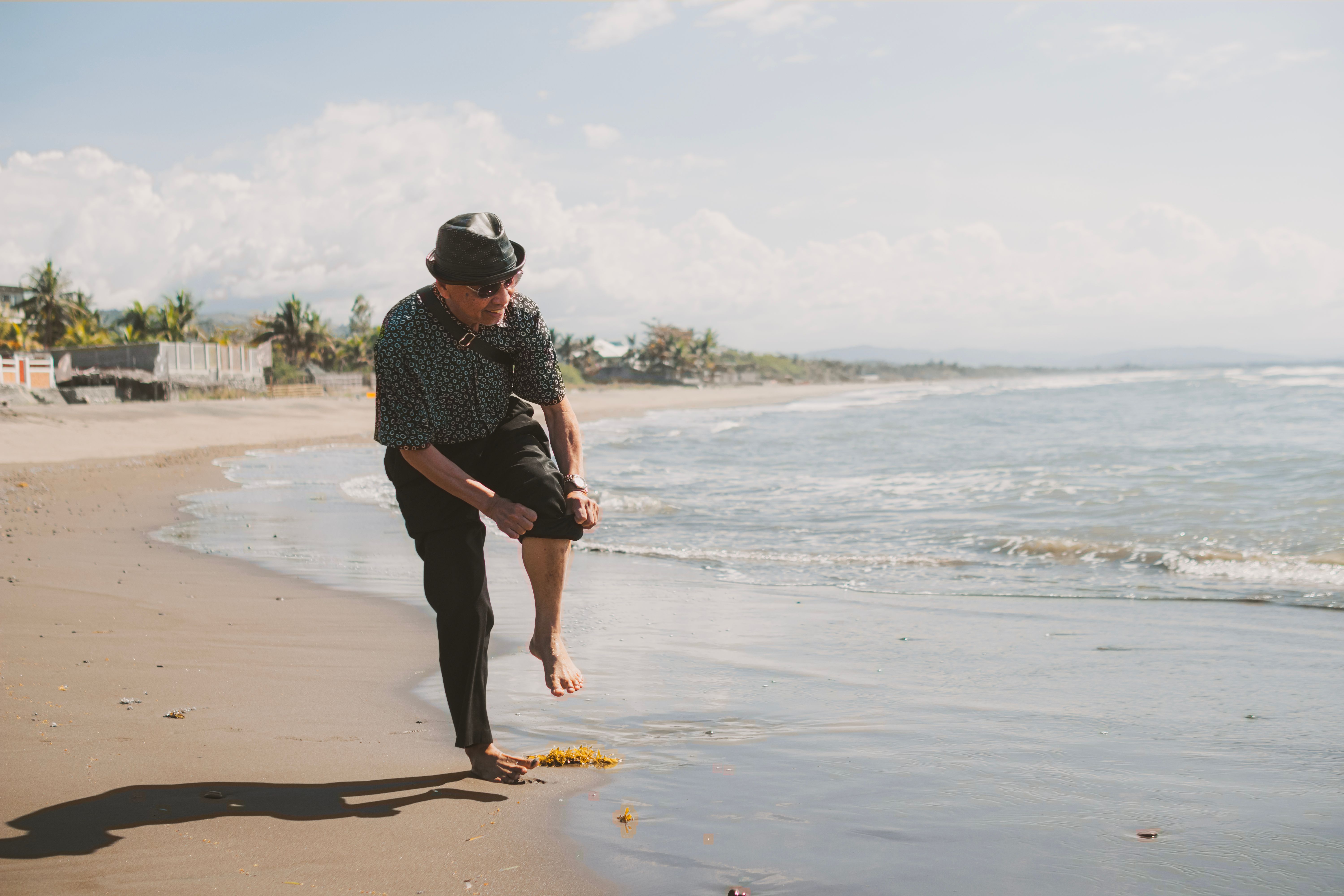 Senior adult man in casual attire walking barefoot on a sunny beach by the sea.