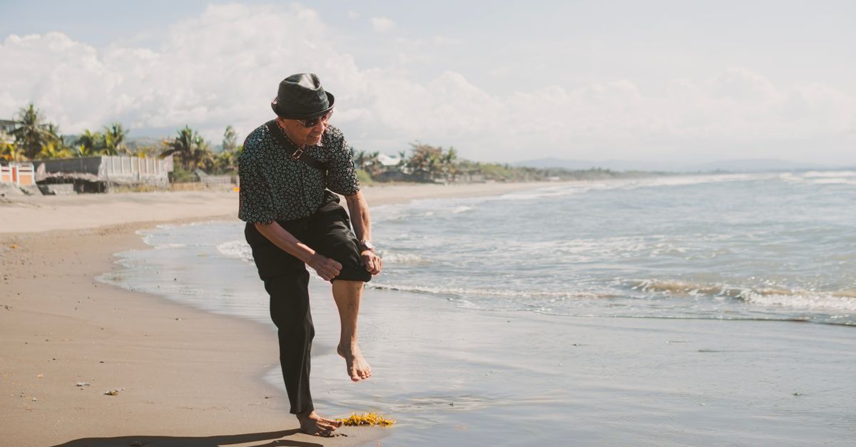 Senior adult man in casual attire walking barefoot on a sunny beach by the sea.