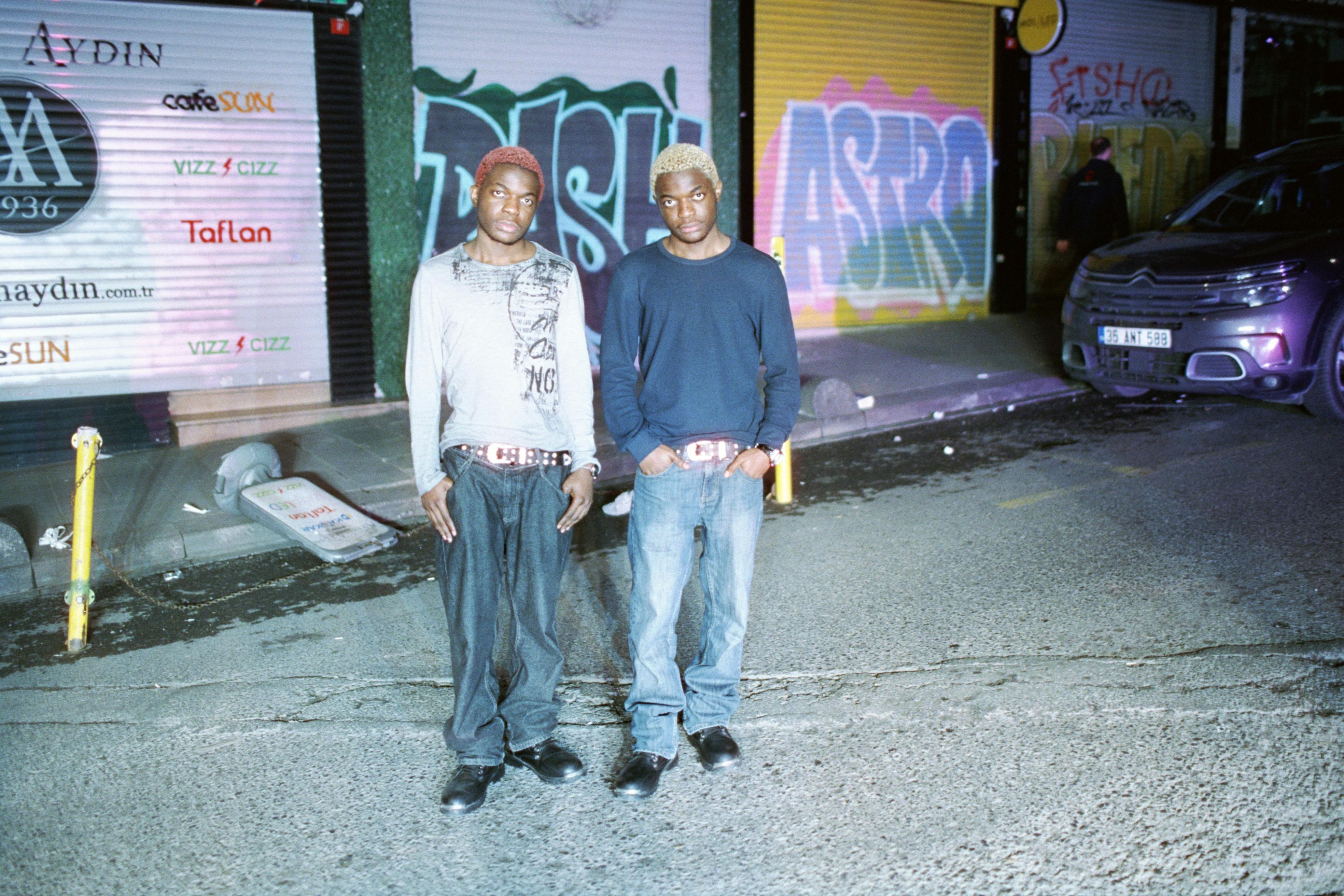 Two men pose on a night-lit urban street with graffiti and storefronts.