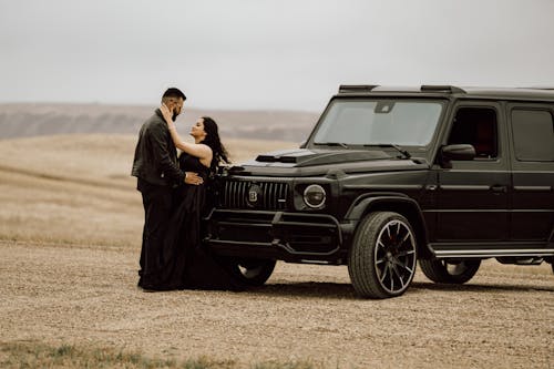 A Couple in Black Clothing Standing next to a Black Car on a Field 