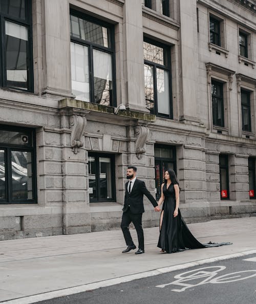 Man in a Suit and Woman in a Black Dress Walking on a Sidewalk in City 