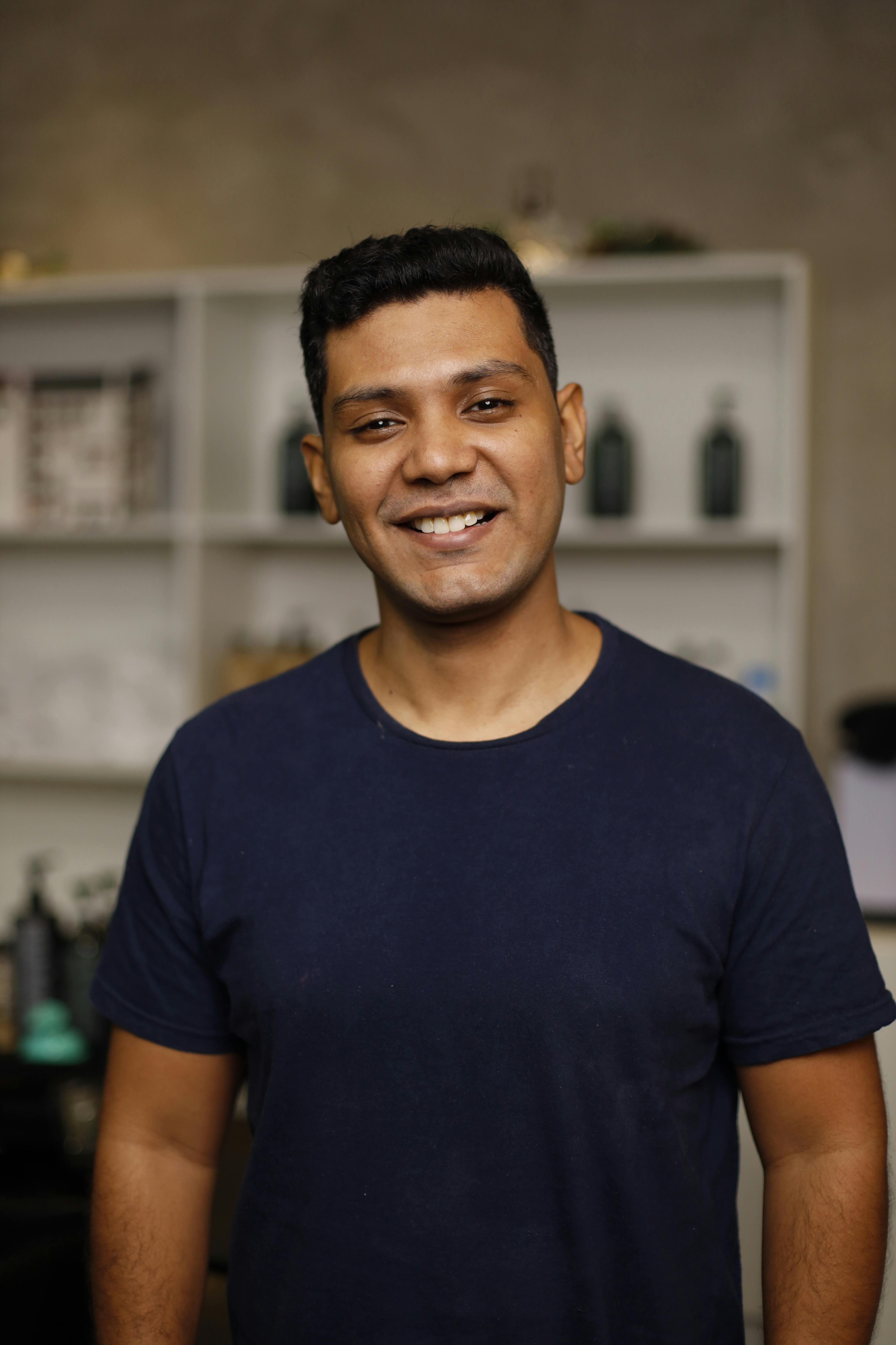 A young man smiling warmly, standing indoors, wearing a navy blue T-shirt.