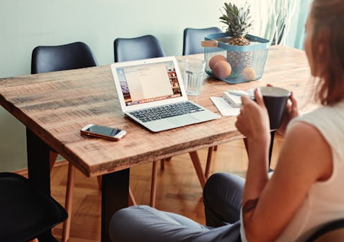 A woman sitting at a wooden table with a laptop, phone, and cup at home, enjoying remote work.