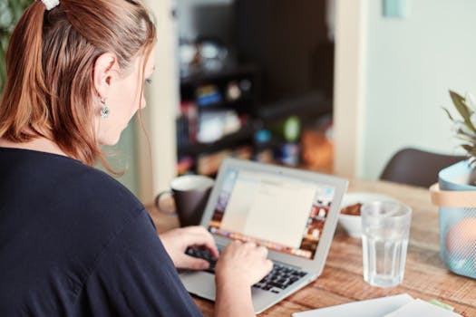 A woman using a laptop at a wooden table in a home office setting with a glass of water and fruit.