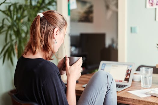 A woman in a cozy home office setting, sipping coffee while working on a laptop.