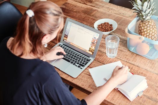 A woman writes notes by her laptop in a cozy dining setting, with fruits and snacks nearby.