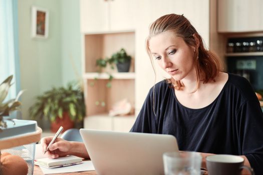 Woman working on laptop in home office, taking notes, surrounded by plants.
