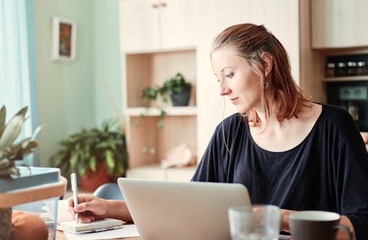 A young woman writes notes while working on a laptop in a cozy home office.