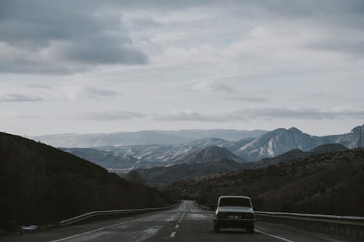 A vintage car travels on a rural mountain road under a cloudy sky, offering scenic serenity.