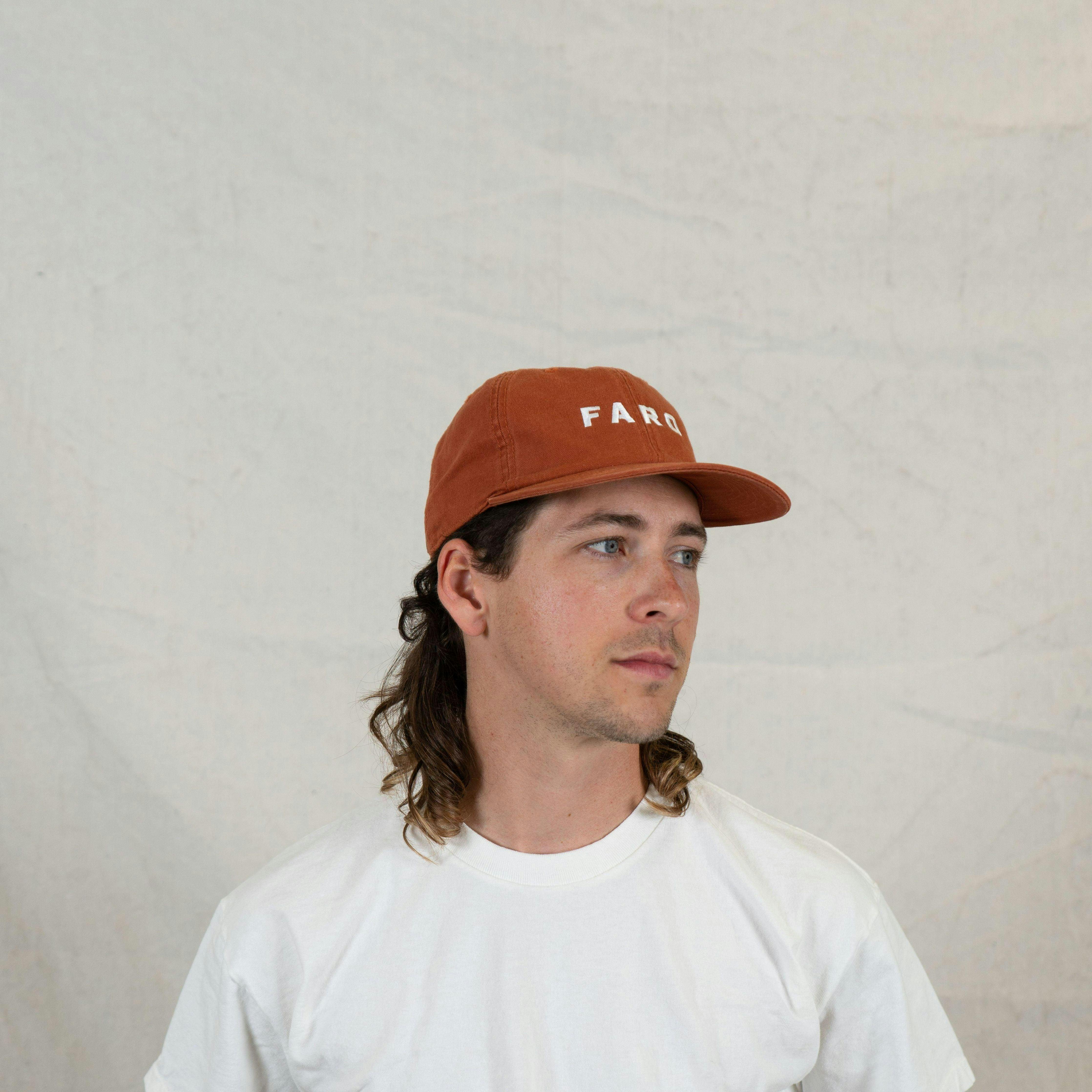 Side profile portrait of a man wearing a brown cap and white t-shirt, set against a neutral background.