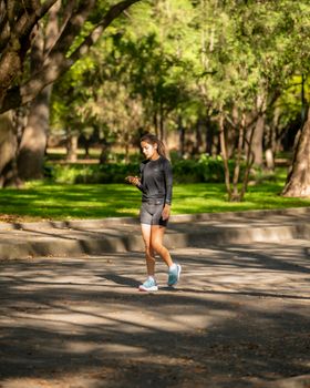 Woman jogging in a sunlit park, checking smartphone while exercising outdoors.