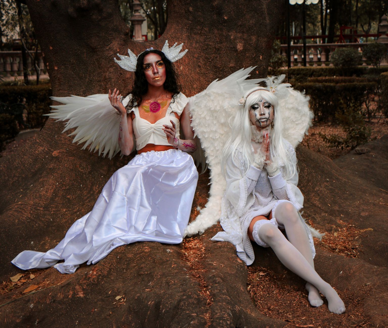 Two women dressed as angels with wings and makeup pose by a tree in a park setting.