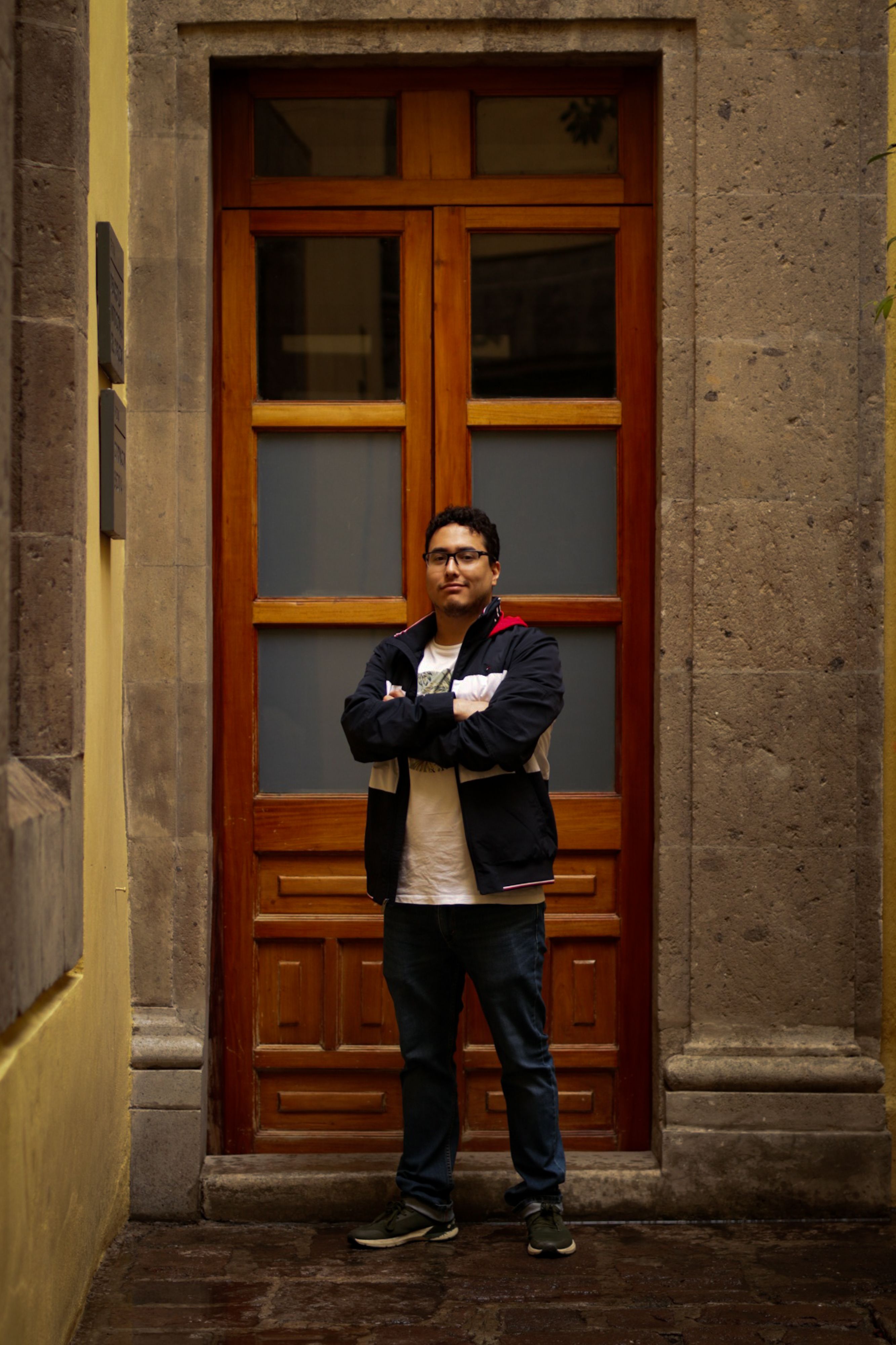 A man stands confidently in front of a wooden doorway in a city street, arms crossed.