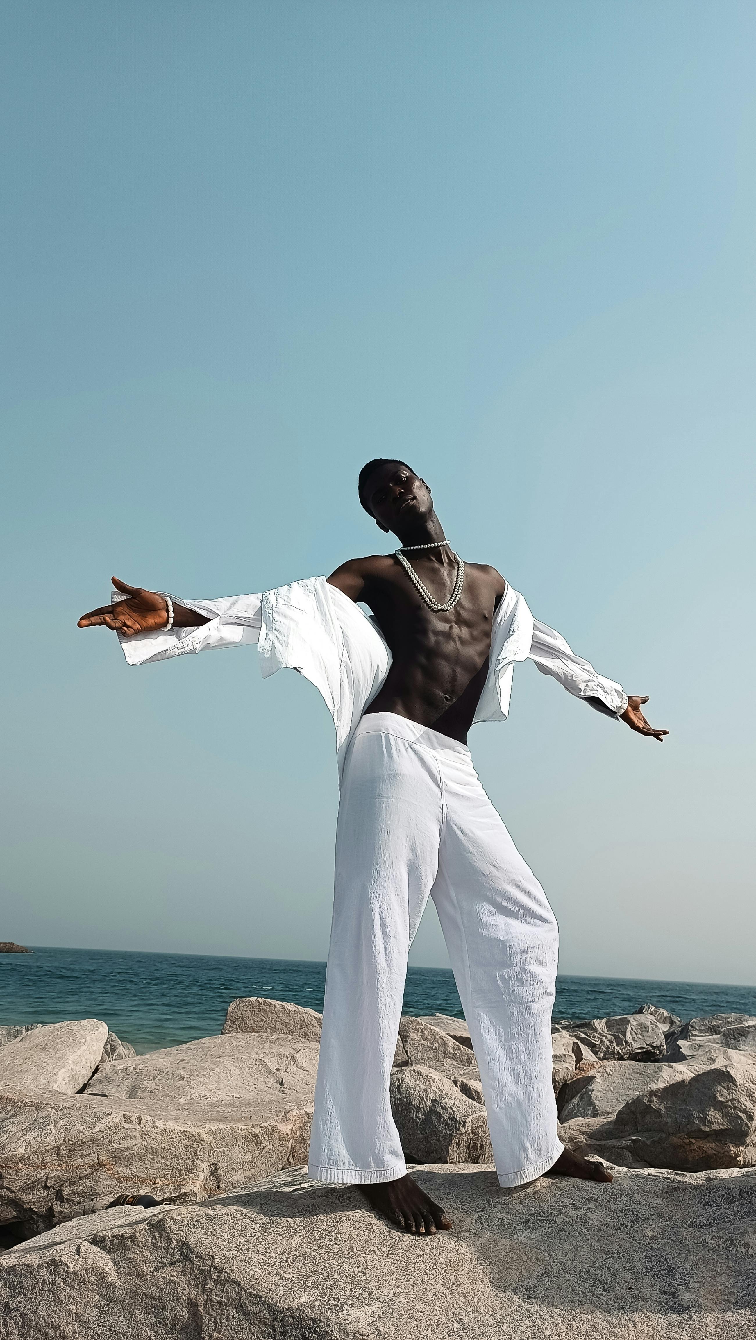 A stylish man in white outfit poses confidently on rocky Lagos coastline under a clear blue sky.