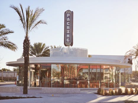 Glass-walled restaurant with palm trees and prominent 'PACKING' sign under clear sky.