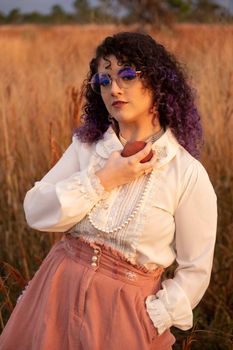 Woman with curly hair and eyeglasses holding an apple in a rural field.